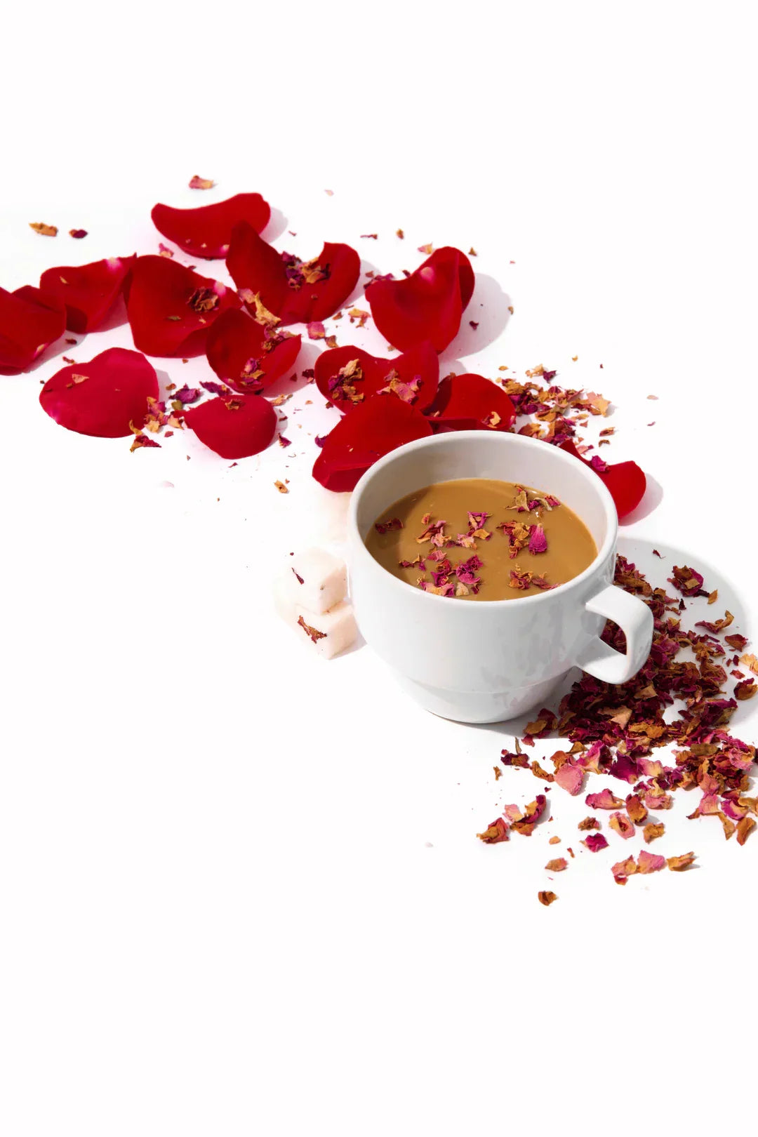 White cup of tea with rose petals on top and scattered around with sugar cubes on white surface