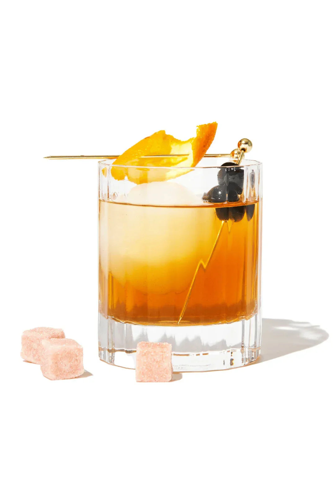 Glass of whiskey cocktail with large ice sphere, orange peel, and dark cherries on gold pick