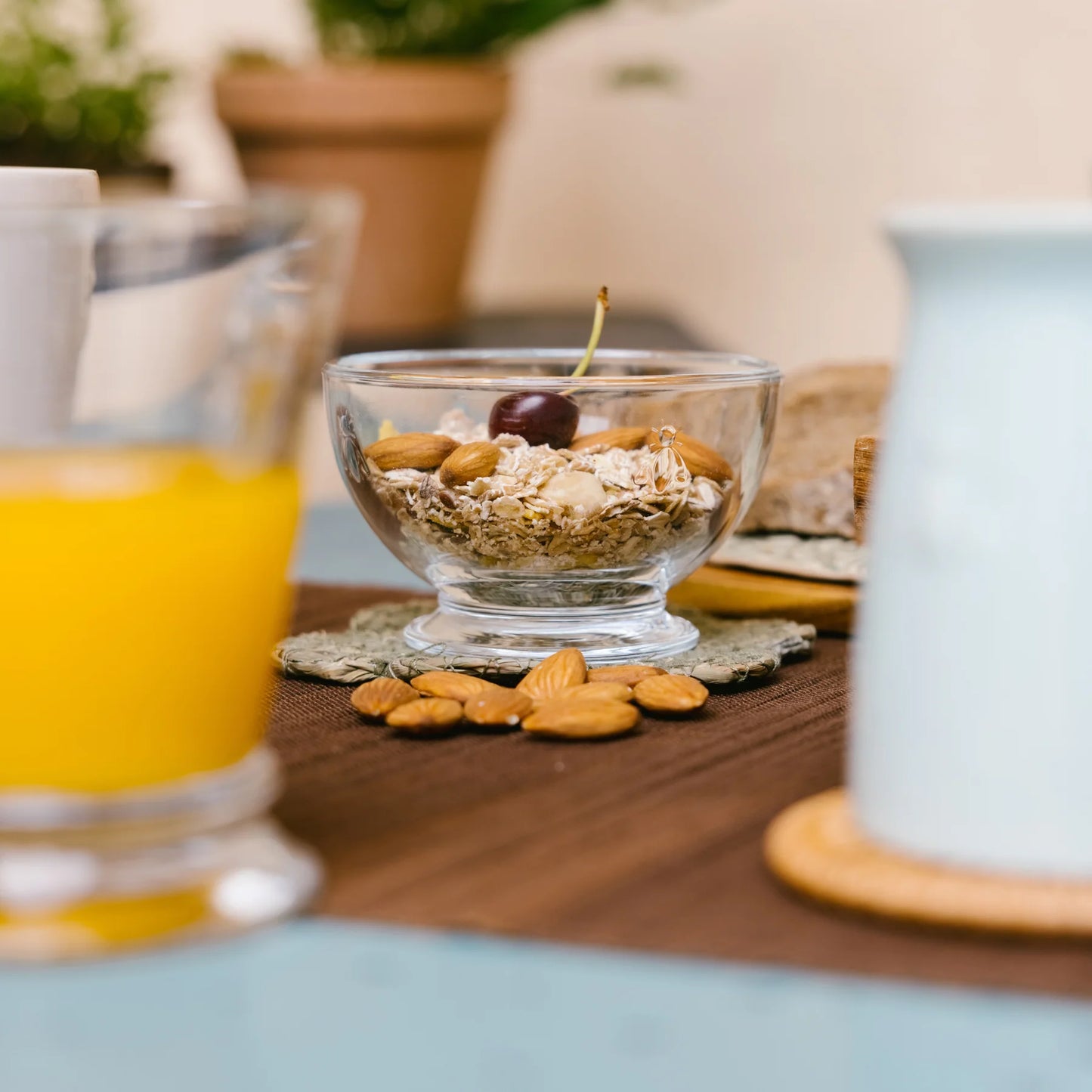 Glass bowl with oats, almonds, and cherry on a table with almonds and blurred juice and milk container