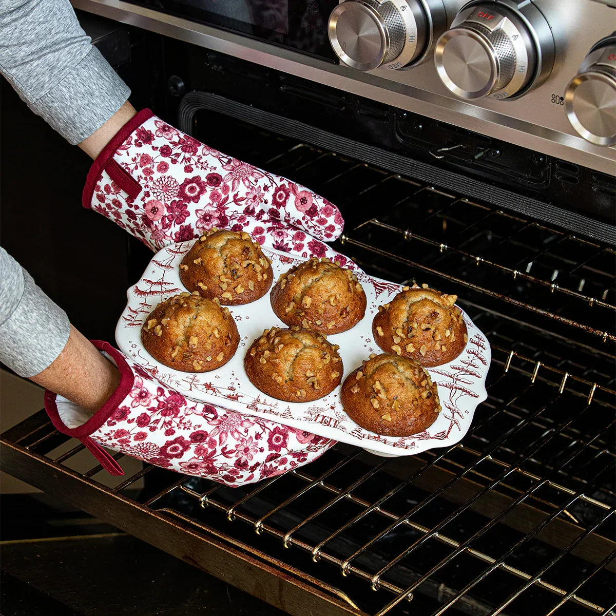 Hands in floral oven mitts holding a ceramic muffin tray with six freshly baked nut-topped muffins by oven