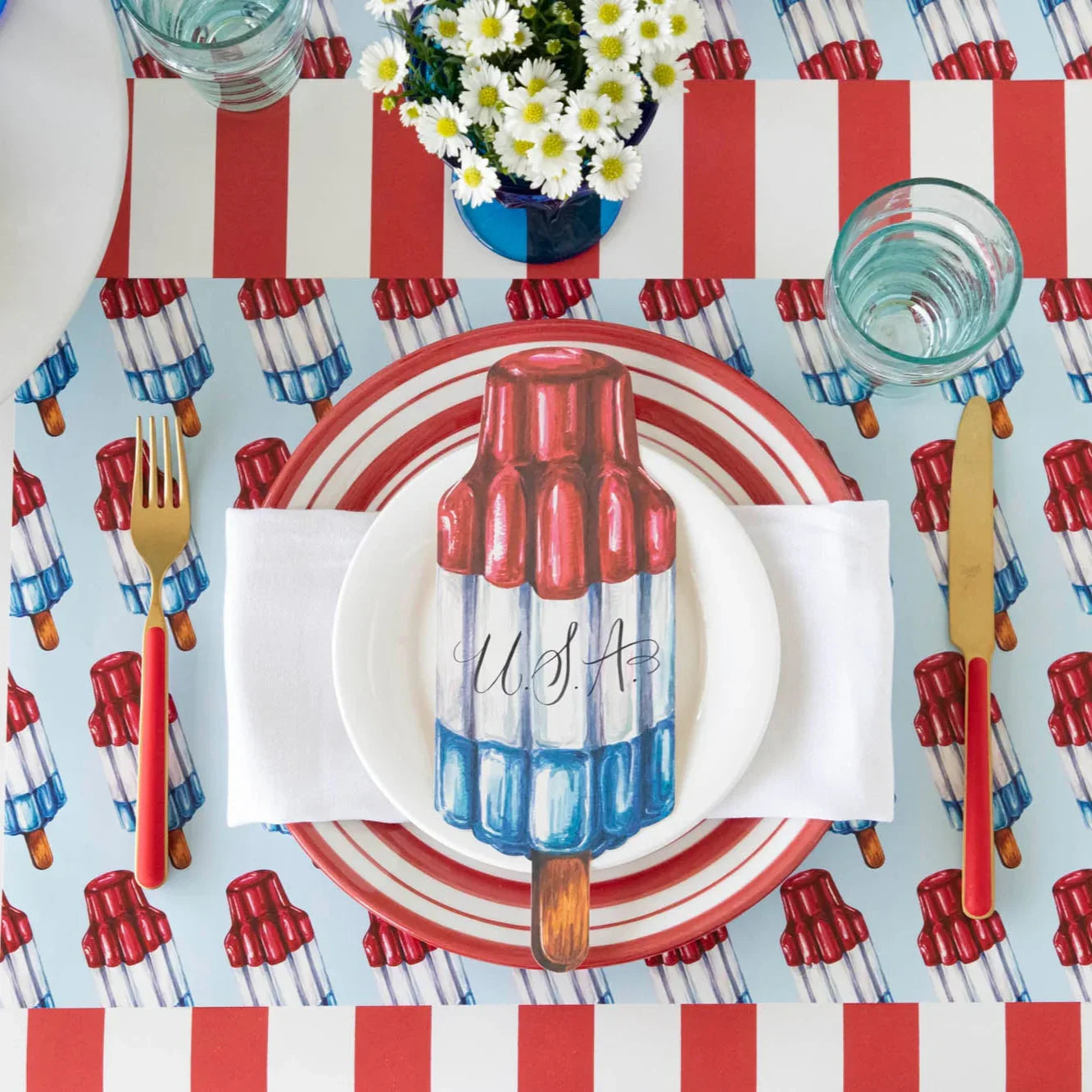 Table setting with USA-themed rocket pop placemat, red-striped plate, gold flatware, and daisies in a blue vase