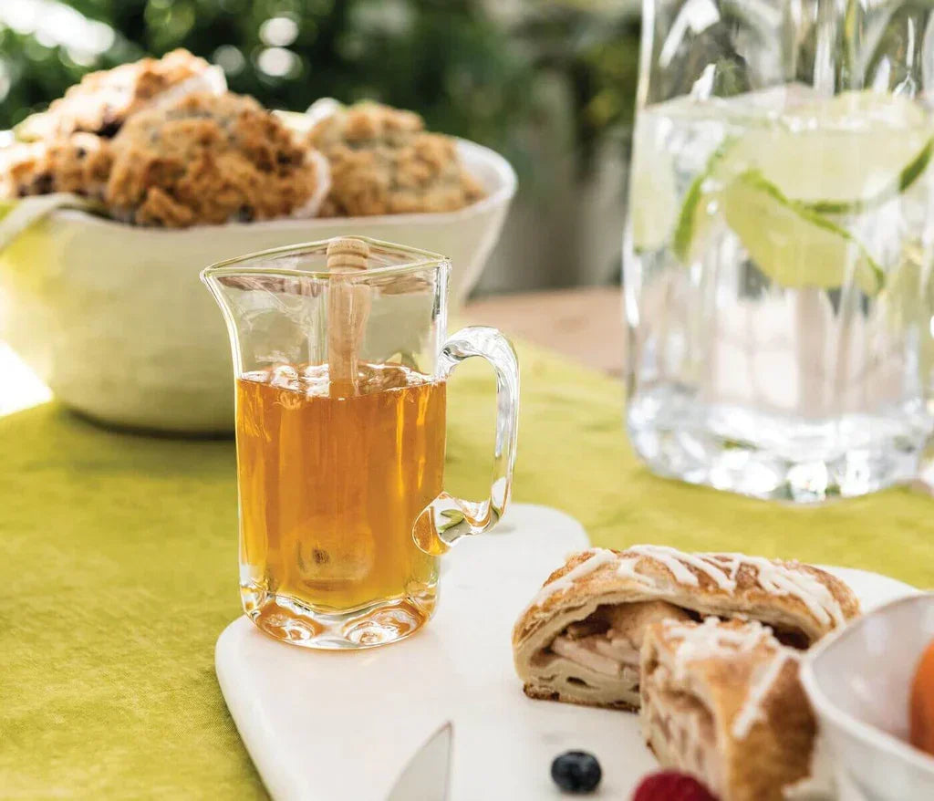 Glass pitcher of honey with dipper, sliced pastry, and fresh berries on brunch table