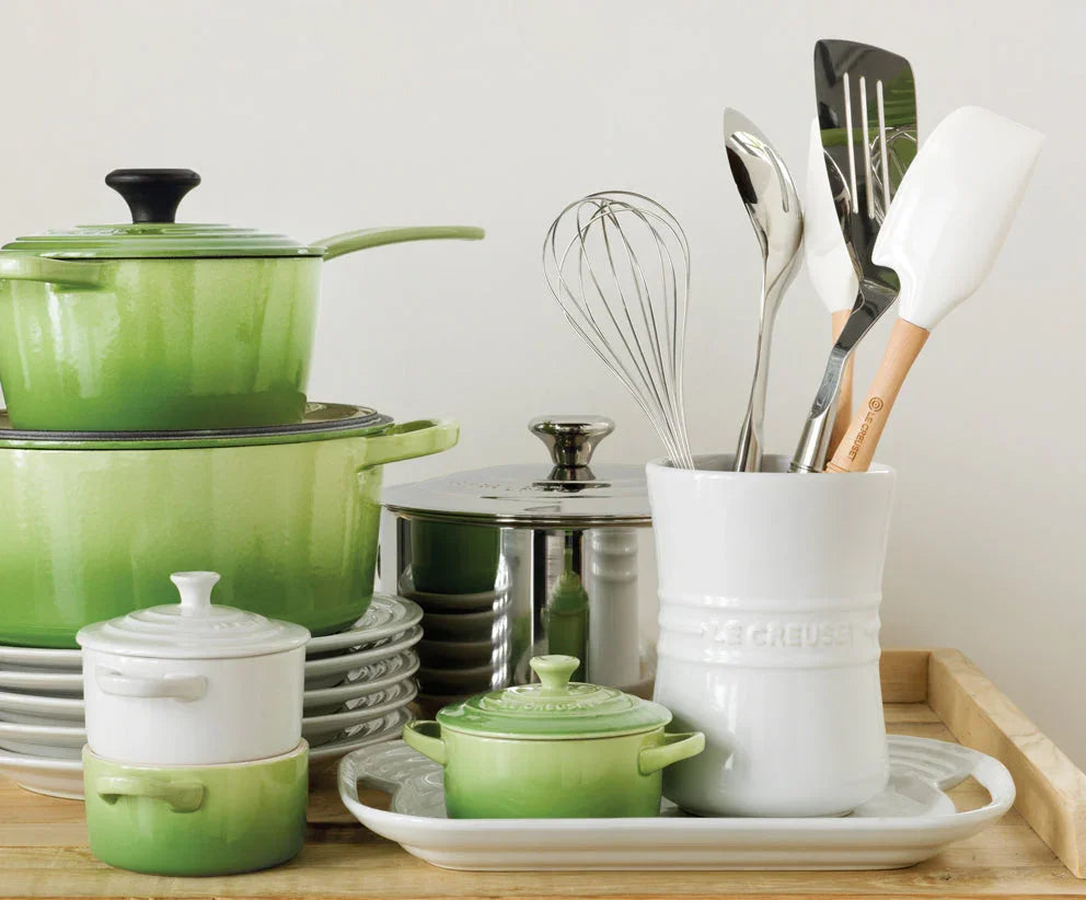 Green and white Le Creuset cookware set with pots, pans, white utensil holder, and kitchen utensils on wooden tray