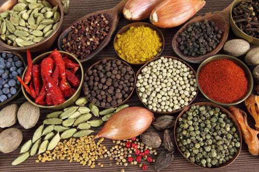 Assorted colorful spices including red chilies, cardamom, white pepper, turmeric, black pepper, and shallots on wooden surface