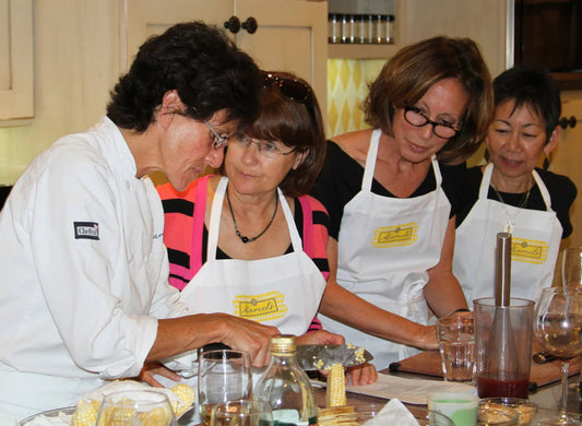 Chef demonstrating corn cutting in a cooking class with three women wearing aprons in a kitchen