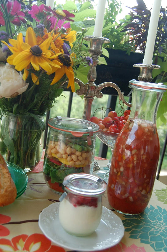 Table with fresh flowers, layered chickpea salad in jar, yogurt dessert, ripe cherries, and a carafe of mixed vegetable drink