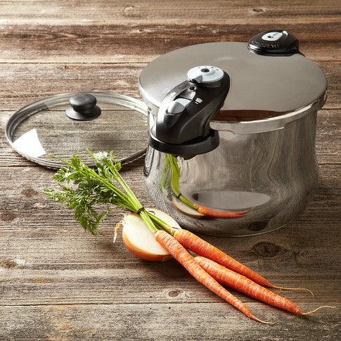 Stainless steel pressure cooker with black handle, glass lid, fresh carrots and onion on wooden surface