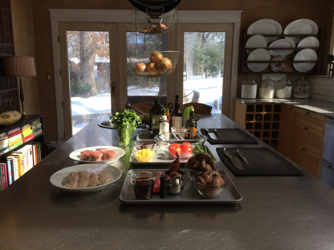 Modern kitchen with cooking ingredients including mushrooms, tomatoes, cilantro, oil bottles, and wrapped chicken on a metal island