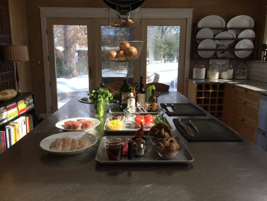 Modern kitchen with cooking ingredients including mushrooms, tomatoes, cilantro, oil bottles, and wrapped chicken on a metal island