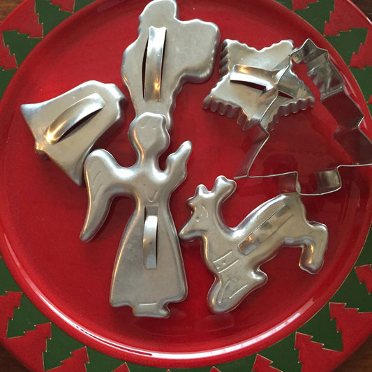 Metal Christmas cookie cutters of an angel, reindeer, star, and bell on a red festive plate with green trees