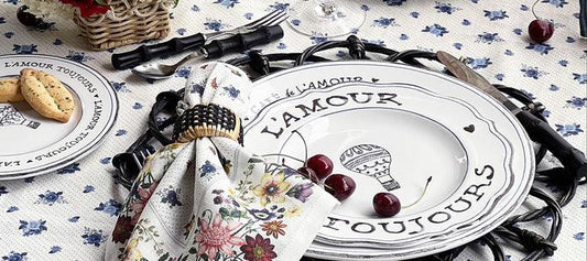 Romantic French-themed table setting with L'Amour Toujours plates, cherries, floral napkin, basket cookies on blue floral tablecloth
