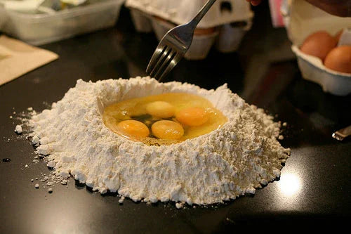 Fresh eggs cracked into a mound of flour on a black countertop for homemade pasta dough preparation