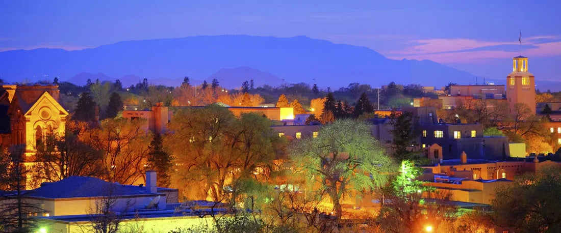 Santa Fe cityscape at dusk with illuminated buildings and distant mountains under a purple sky