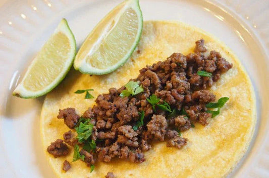 Ground beef taco on corn tortilla garnished with cilantro and lime wedges on white plate