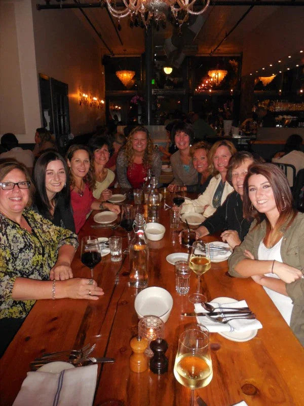 Group of women dining together at a long wooden community table with wine glasses and warm restaurant lighting