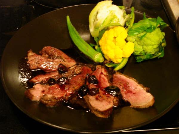 Sliced duck breast with dark sauce and berries, served with green, yellow, and white cauliflower and okra on black plate