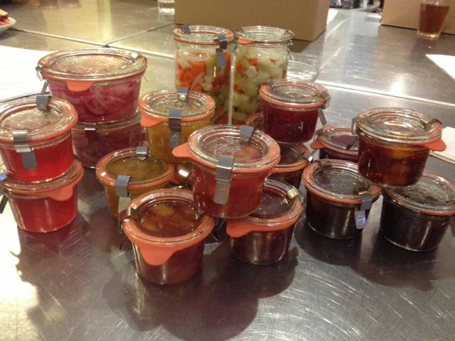 Glass jars filled with various preserved pickled vegetables and sauces on a metal countertop