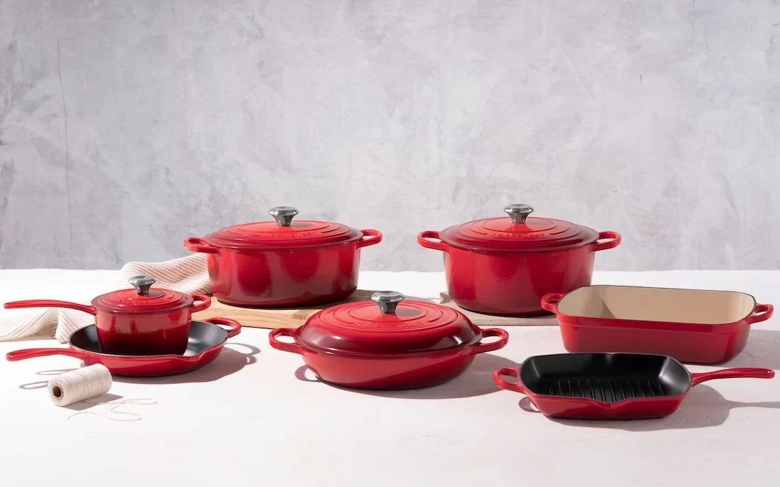 Set of red enameled cast iron cookware including pots, pans, skillet, and grill pan on white surface