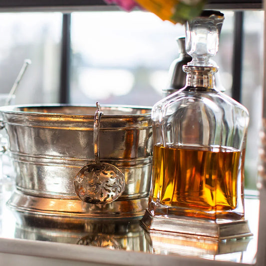 Glass whiskey decanter with amber liquid beside vintage silver ice bucket on reflective surface
