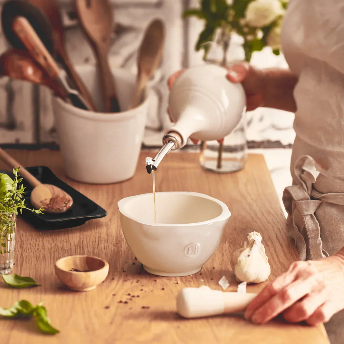 Hand pouring oil into ceramic mortar on wooden kitchen counter with herbs, garlic, and wooden utensils