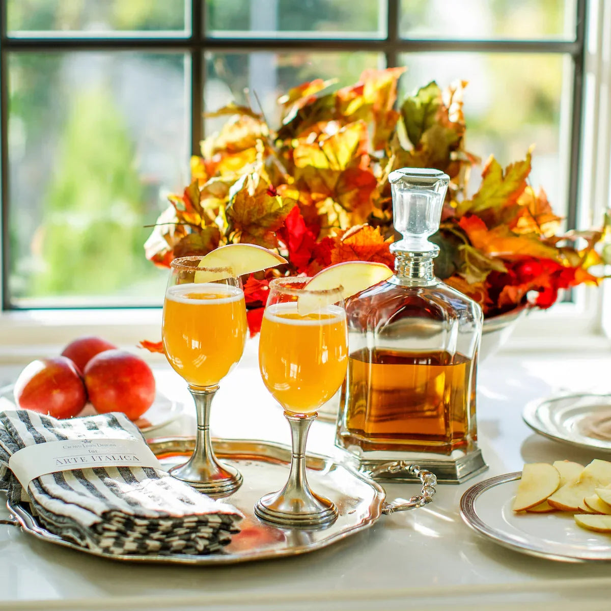 Two apple cider cocktails with apple slices and a glass decanter on silver tray by autumn leaves