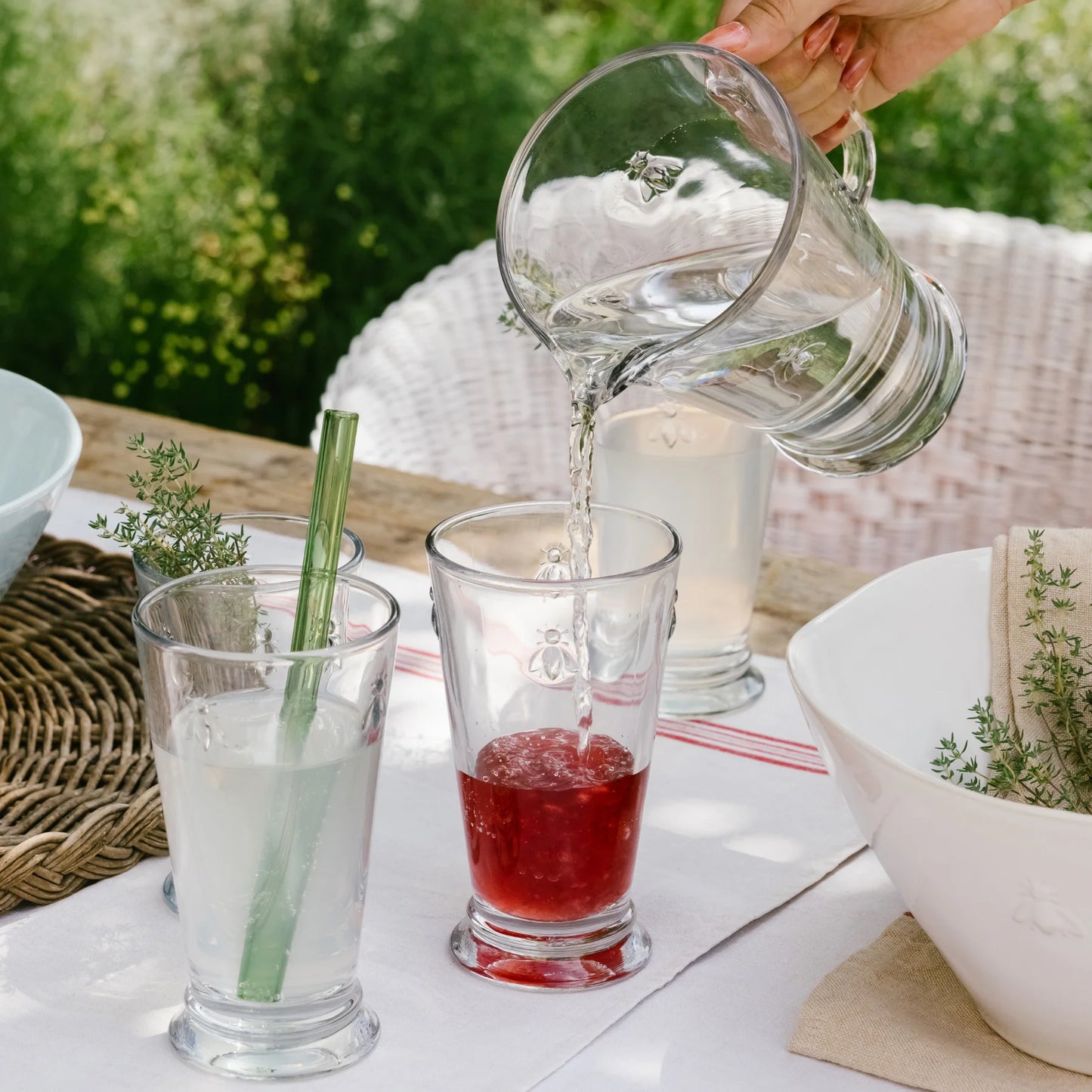 Hand pouring water into glass with red liquid, refreshing outdoor drink with greenery