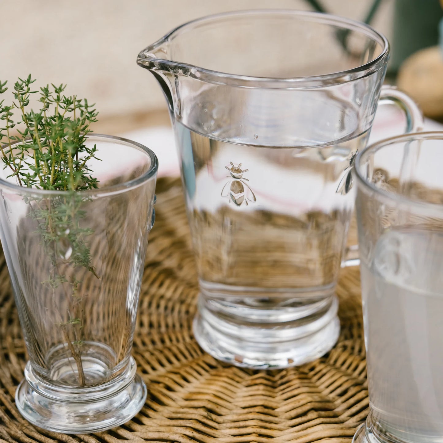 Glass pitcher and glasses with bee design on wicker tray, one glass with green herb sprig