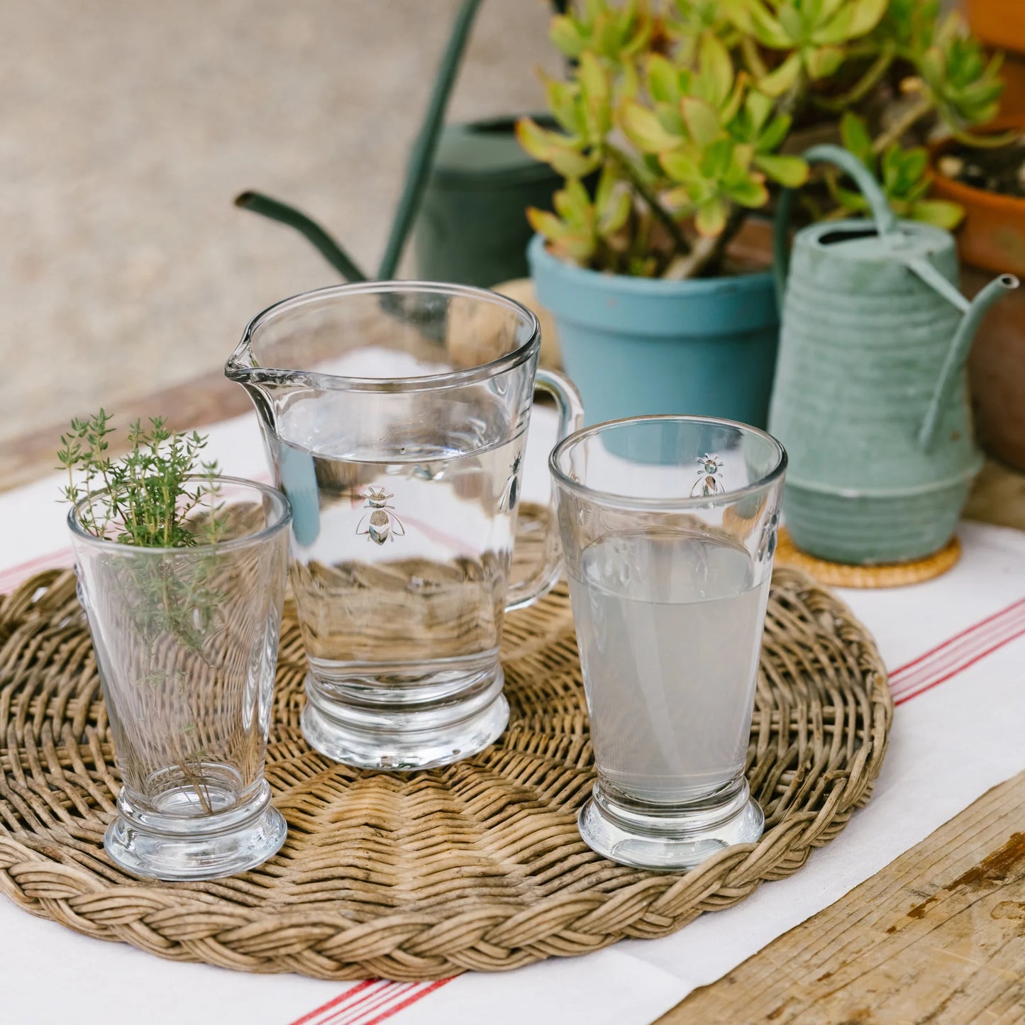 Wicker tray with glass pitcher, two glasses with herbal water and plant sprig on white tablecloth