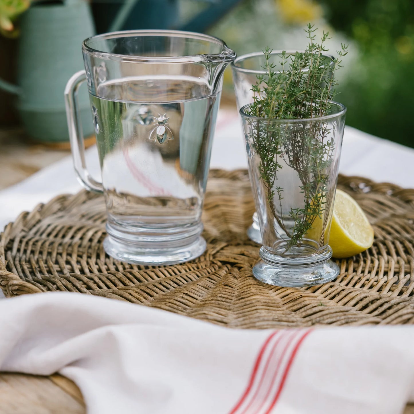Glass pitcher of water and glass with fresh thyme on wicker tray with lemon wedge