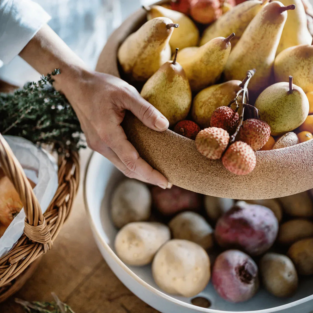 Hand holding bowl of pears and lychee fruit above basket of potatoes and plums on wooden table