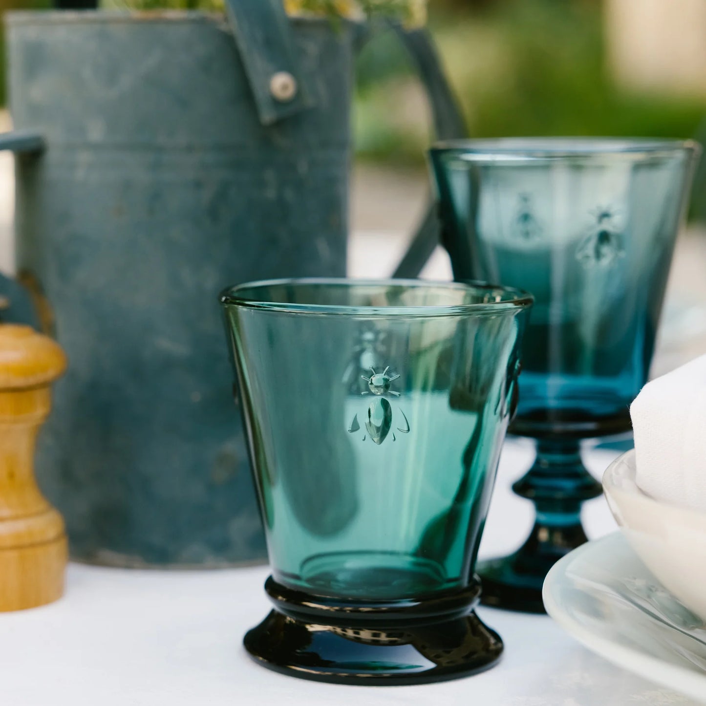 Two blue-green embossed glass cups with bee designs on a white surface, rustic metal pitcher and wooden pepper grinder