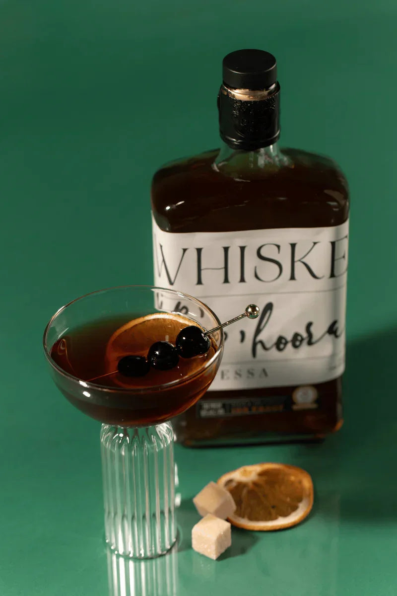 Whiskey cocktail with cherries and orange slice in glass next to whiskey bottle and sugar cubes