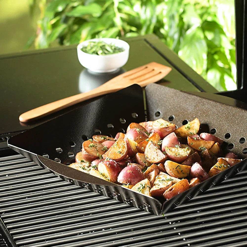 Grill basket with seasoned red potatoes cooking on outdoor grill with wooden spatula and herb bowl