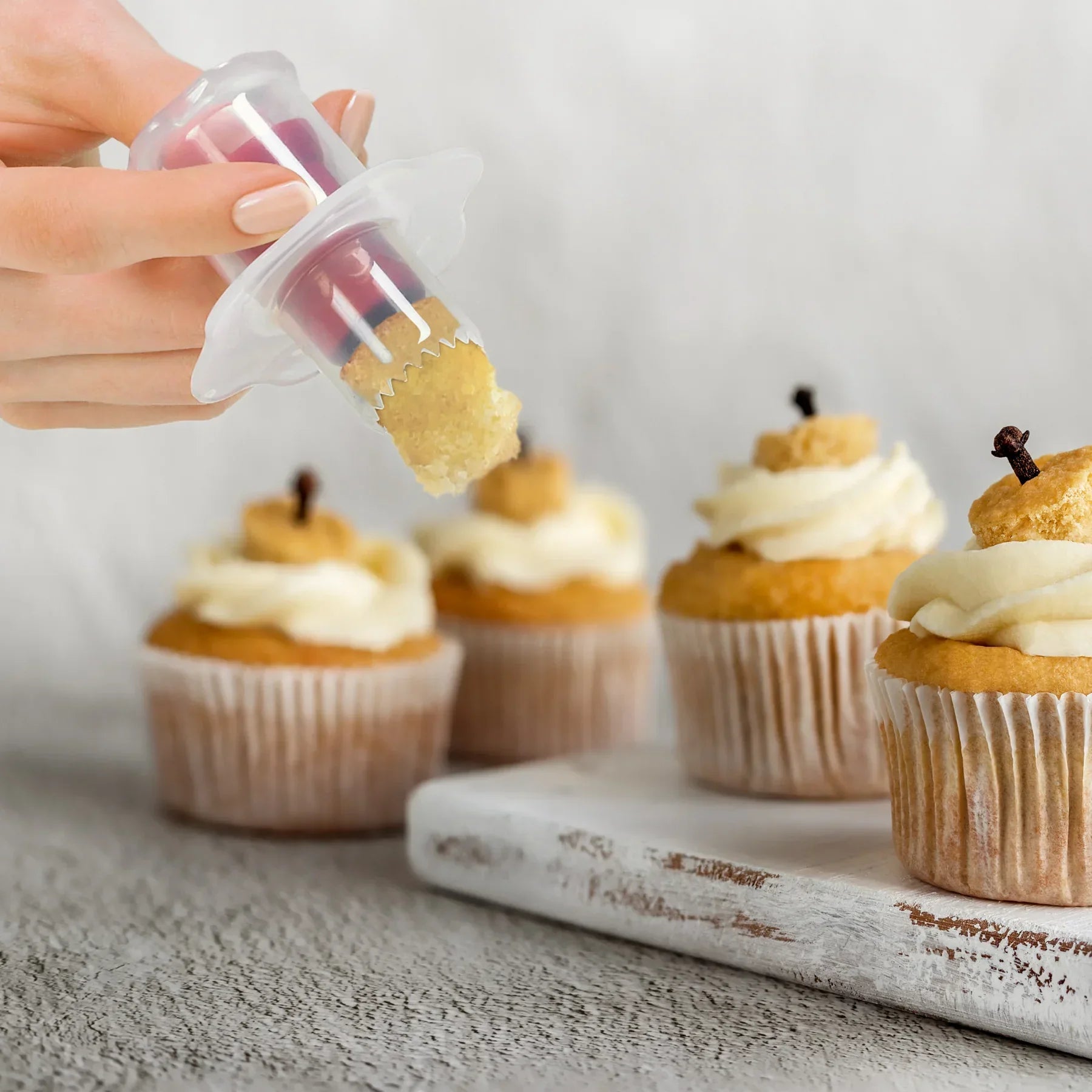 Hand holding cupcake corer tool removing core from vanilla cupcake with frosting on wooden table