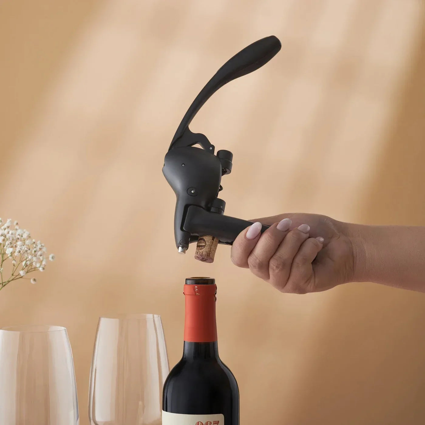 Hand using black lever corkscrew to open red wine bottle on beige background with wine glasses