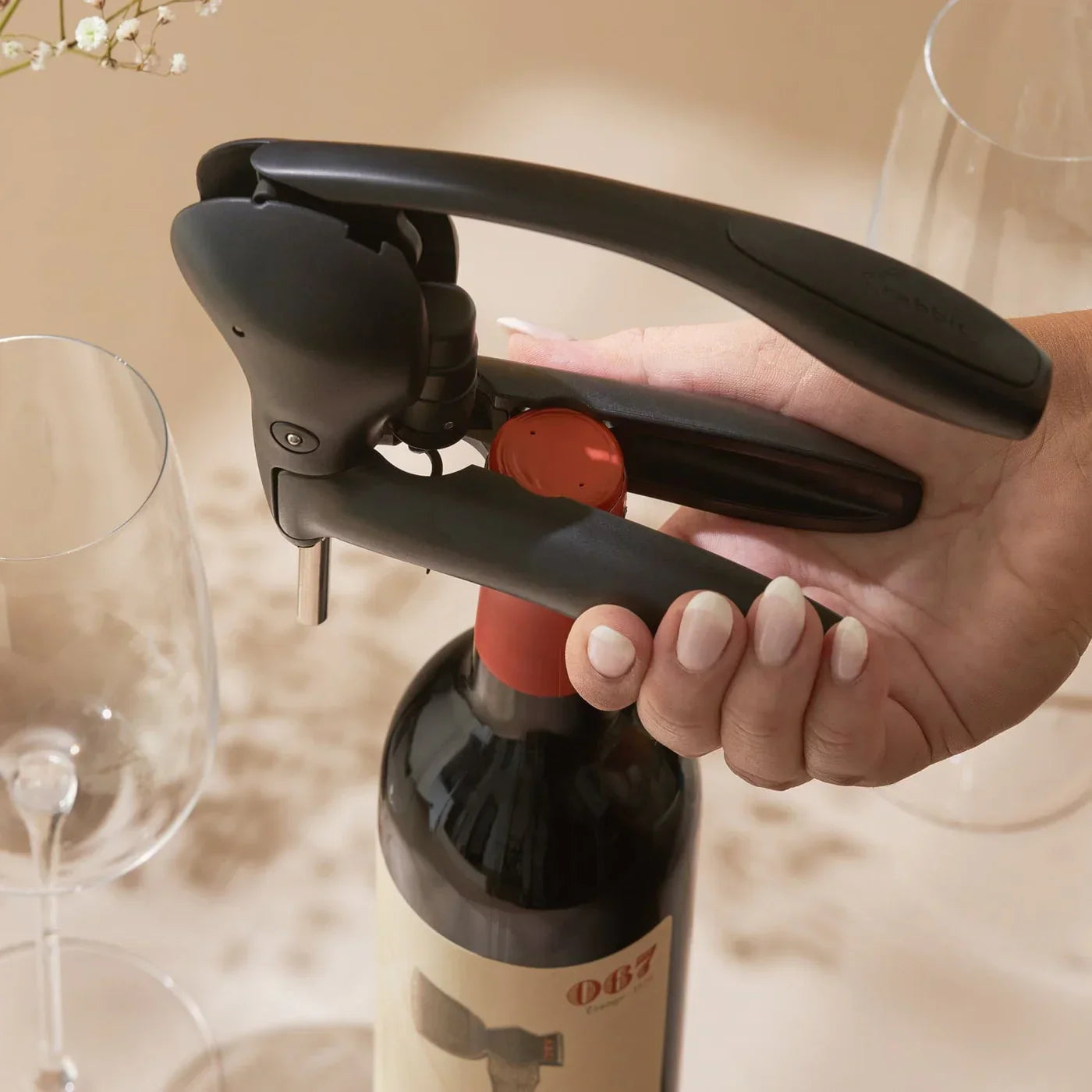 Hand using black lever corkscrew to open red-capped wine bottle on beige table with wine glasses