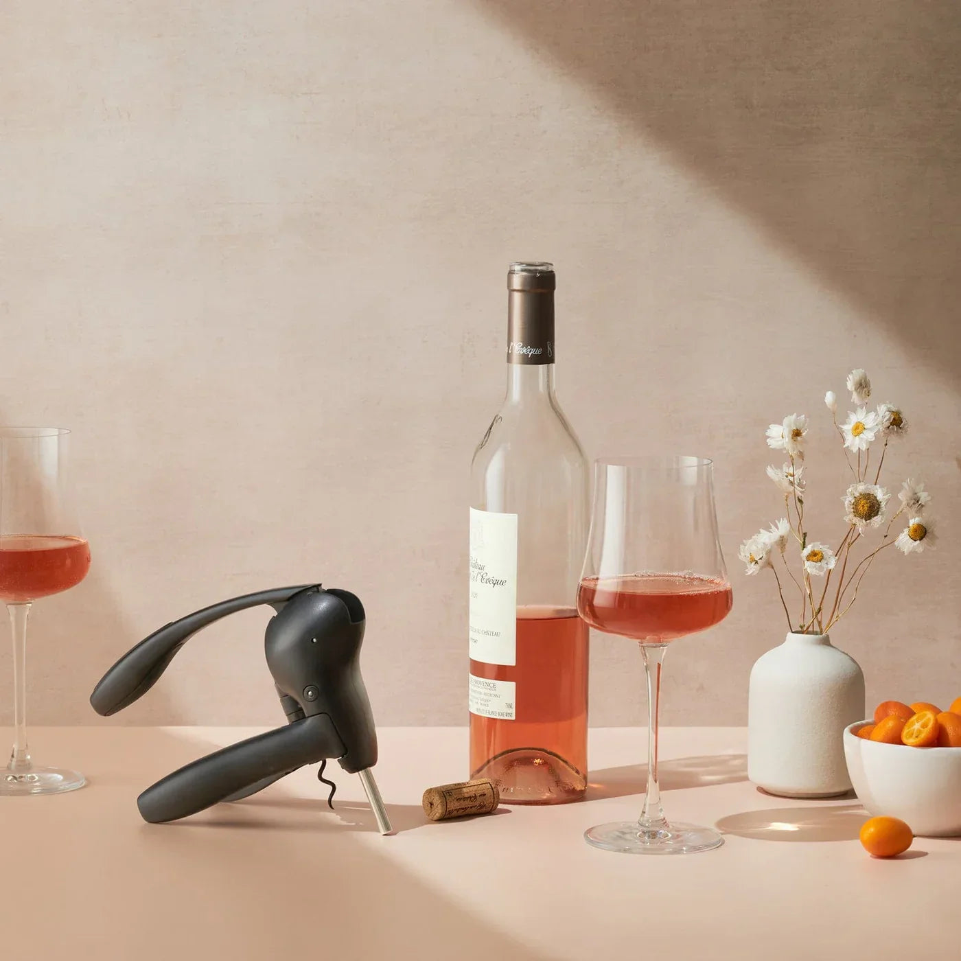 Black lever corkscrew opening rosé wine bottle near filled wine glass, small vase with daisies, and bowl of kumquats
