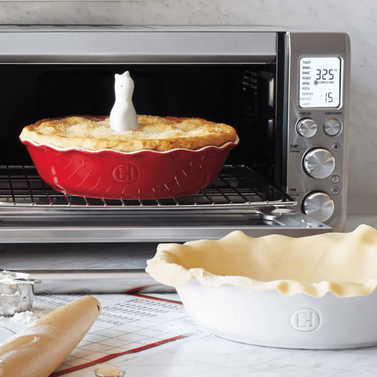 Red pie dish with baked pie and ceramic bird vent in toaster oven, unbaked pie crust in white dish nearby