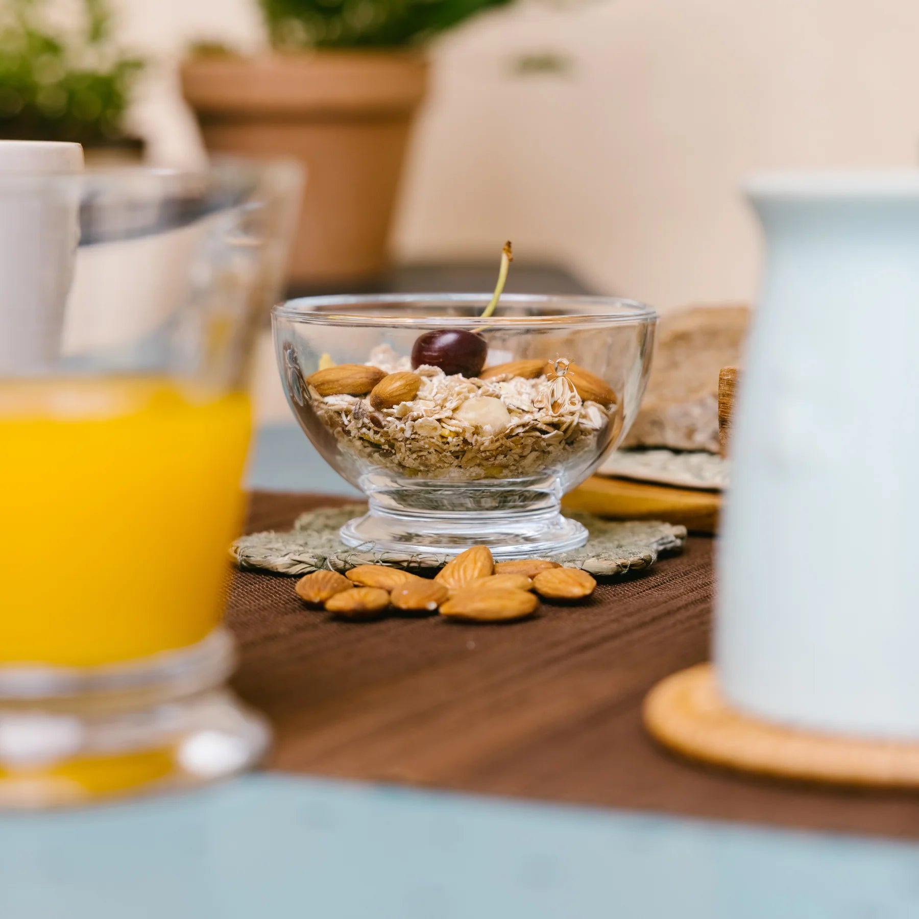Glass bowl with oats, almonds, and cherry on a table with almonds and blurred juice and milk container