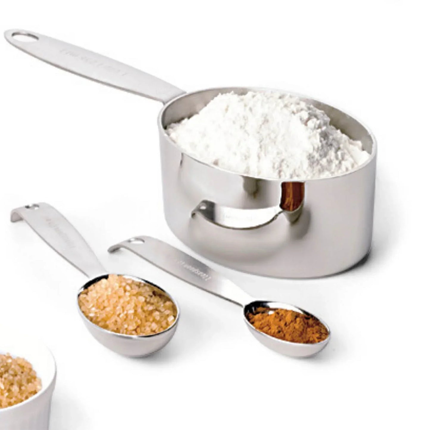 Stainless steel measuring cup with flour, measuring spoons with brown sugar and cinnamon on white background