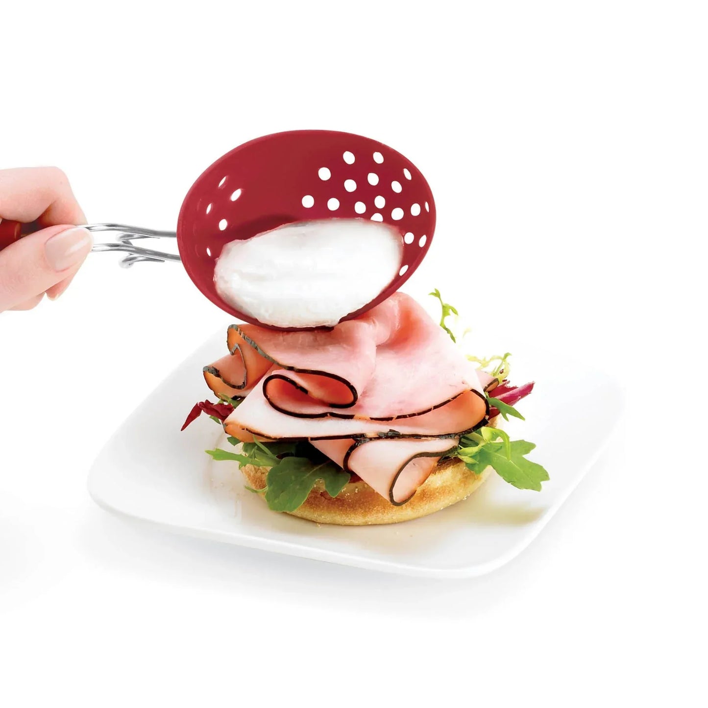 Hand holding red slotted spoon with poached egg over sandwich with sliced turkey and greens on white plate