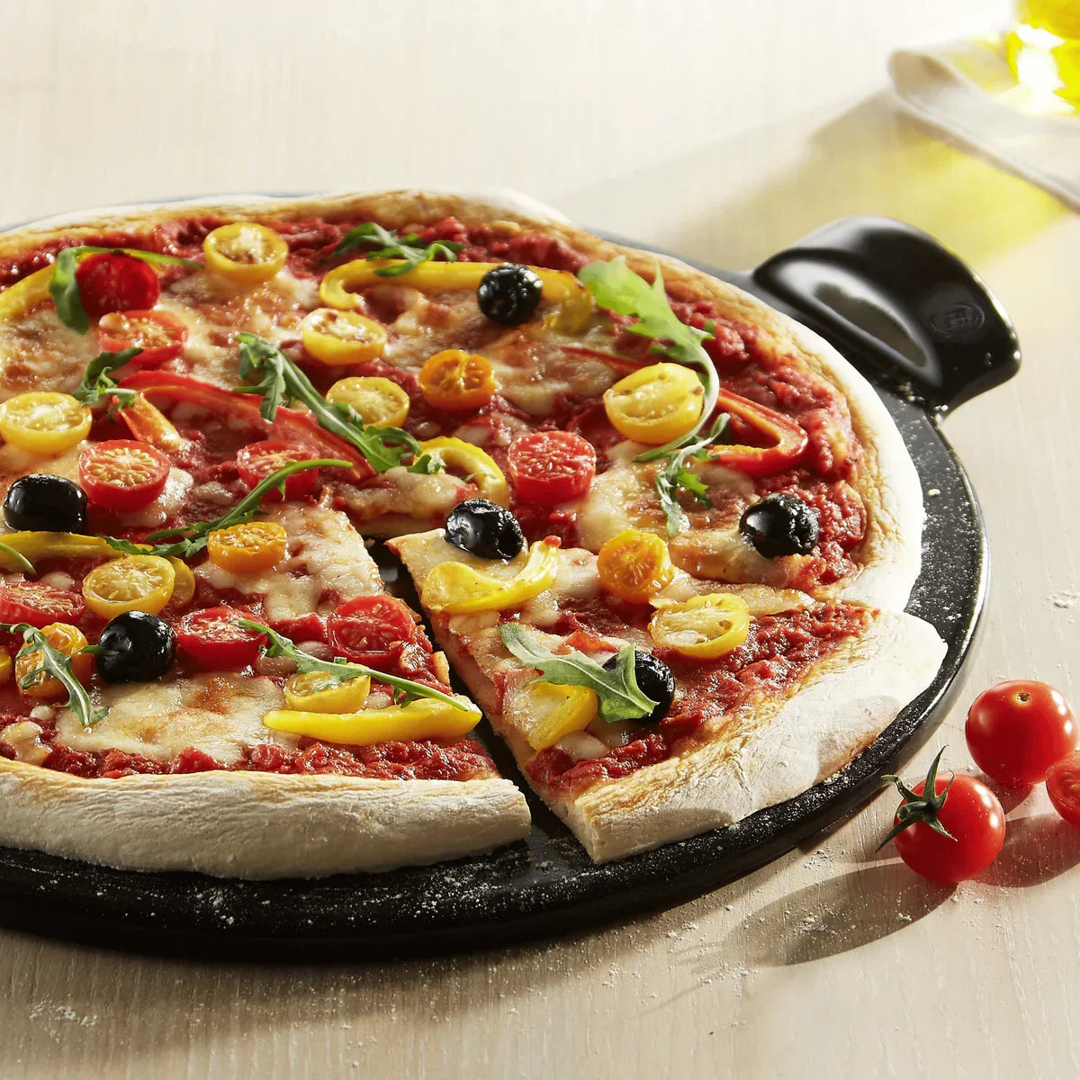 Vegetarian pizza with yellow and red cherry tomatoes, black olives, and arugula on a charcoal pizza stone