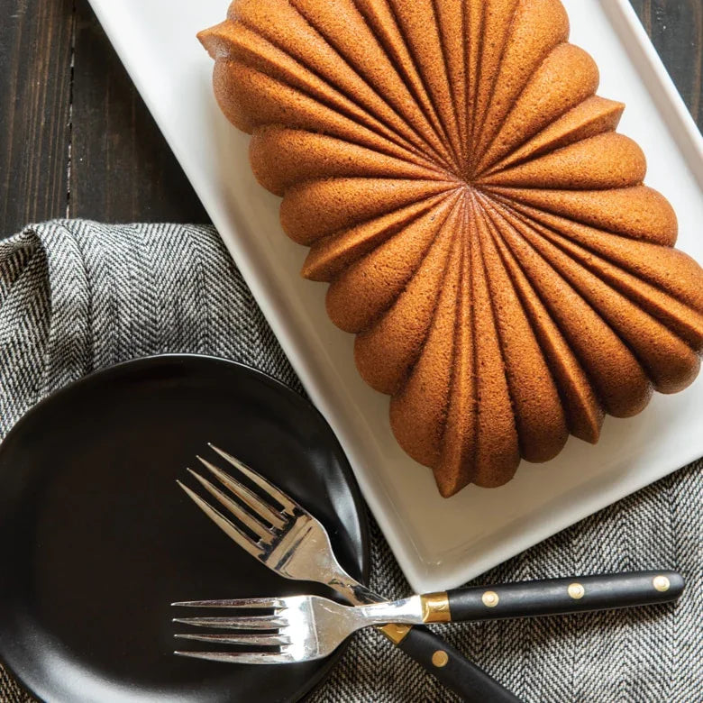 Golden brown classic fluted loaf cake on white platter with black plate and forks on grey cloth