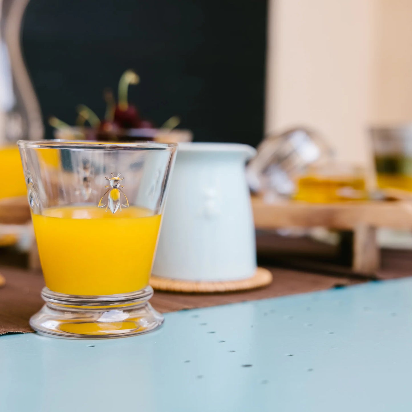 Glass of orange juice with bee emblem on blue table and blurred background jug and cherries