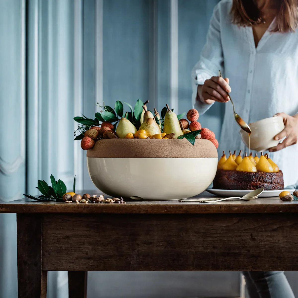 Large cream and clay storage bowl filled with pears, lychees, and greenery on wooden table
