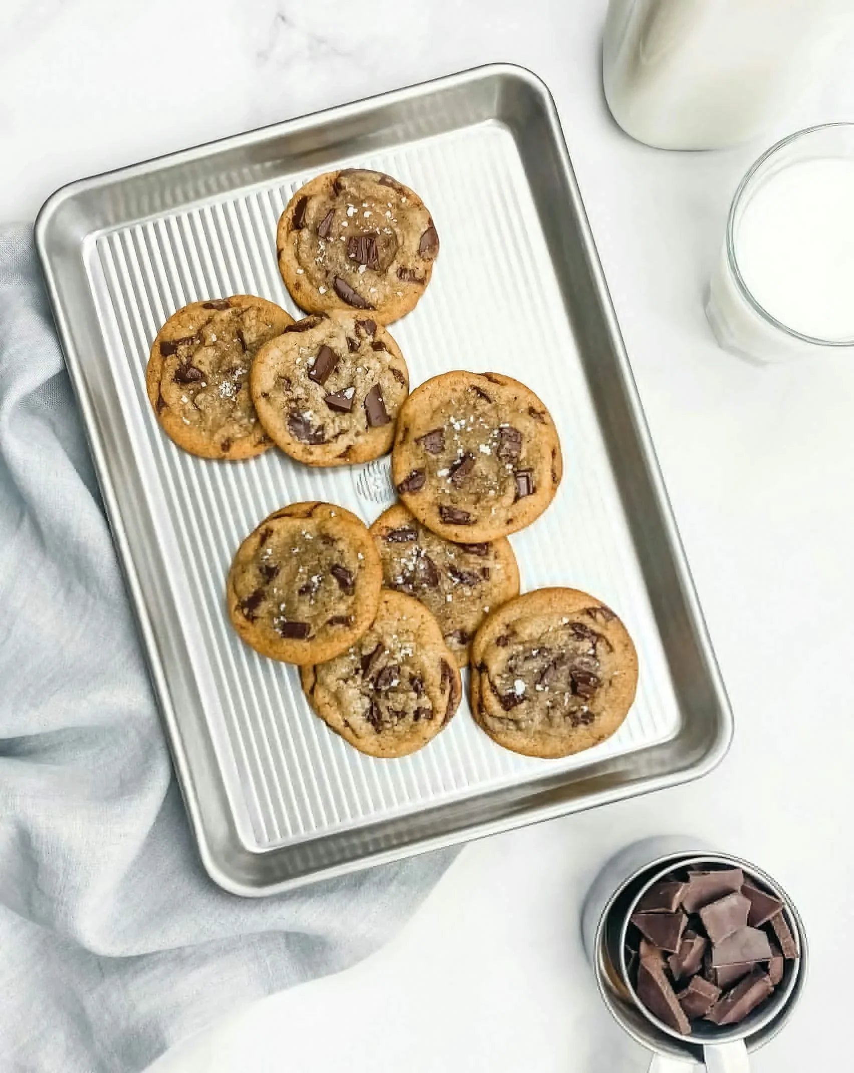 Tray of seven chocolate chunk cookies sprinkled with sea salt on a metal baking sheet with milk and chocolate pieces