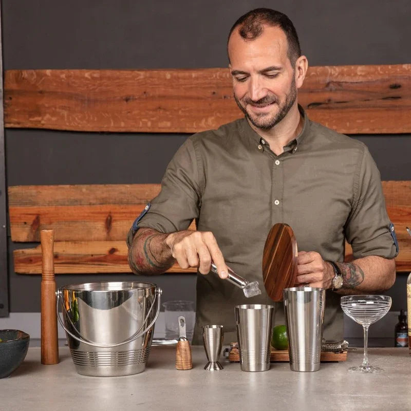 Man with tattoos preparing cocktail using ice cubes, metal shaker, and wooden bar tools on counter