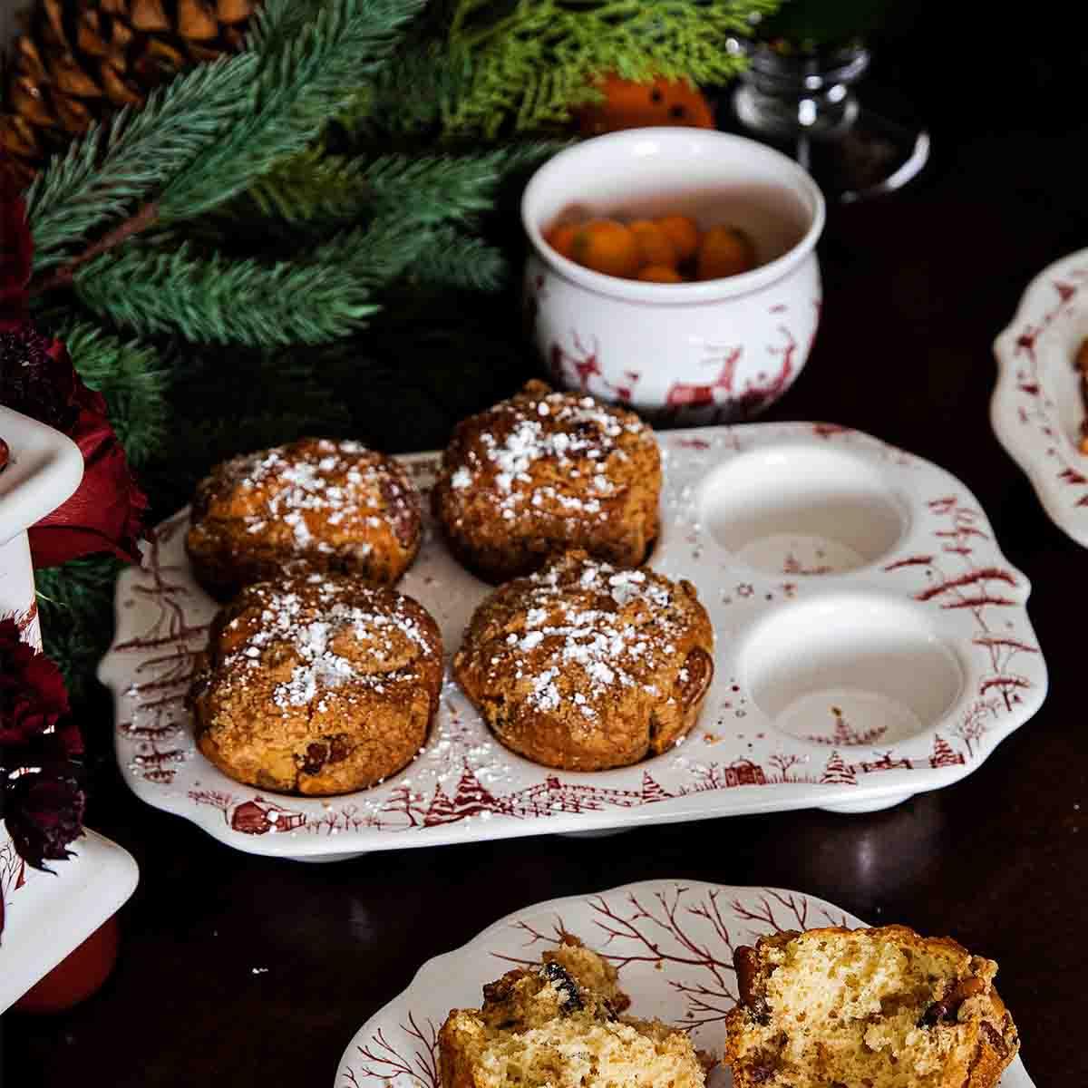 Four powdered sugar-dusted muffins on a decorative holiday ceramic tray with pine greenery and a cup of berries nearby