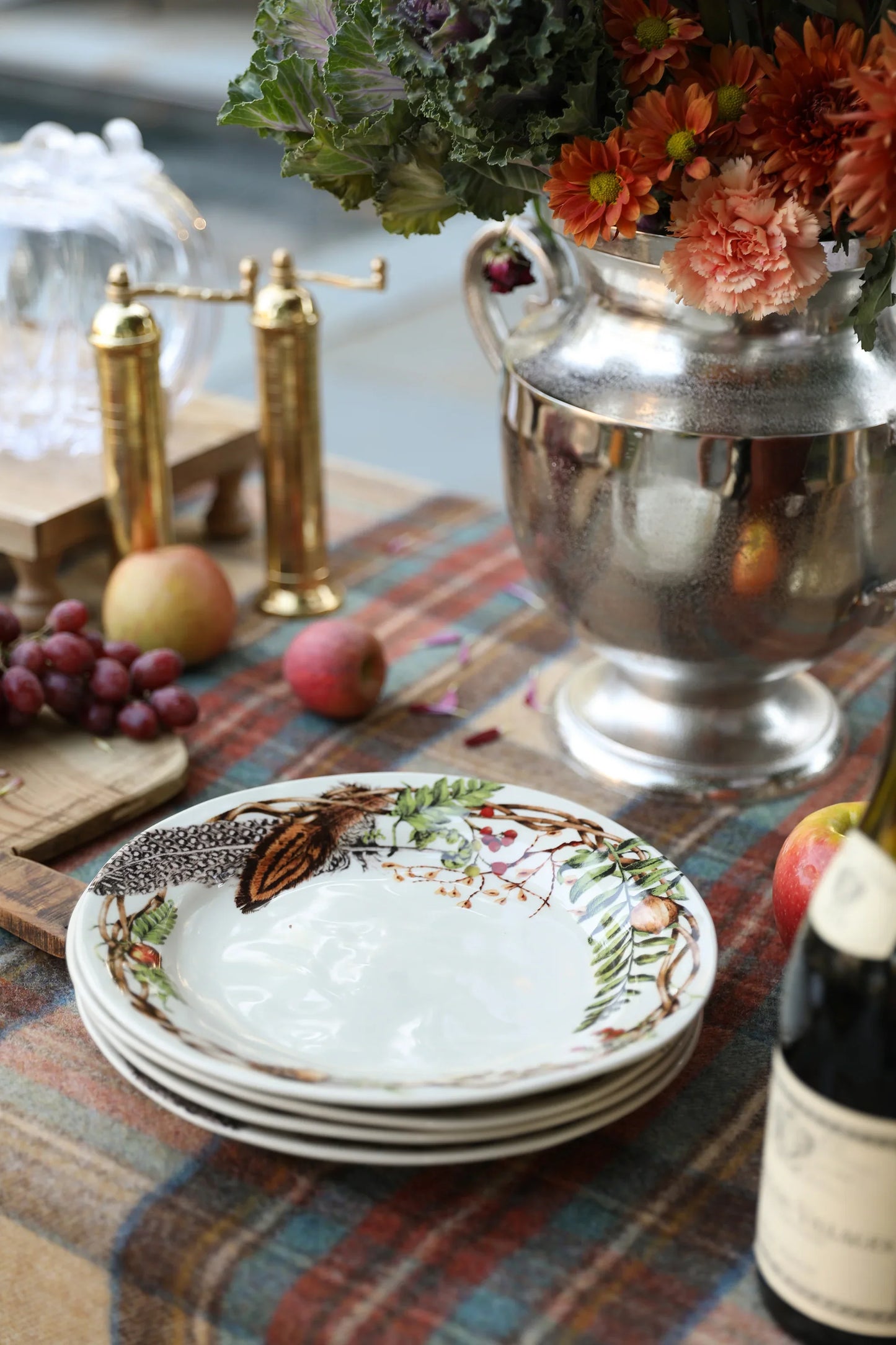 Stack of decorative woodland-themed plates on a plaid tablecloth with brass pepper mills, apples, grapes, and a silver vase with autumn flowers