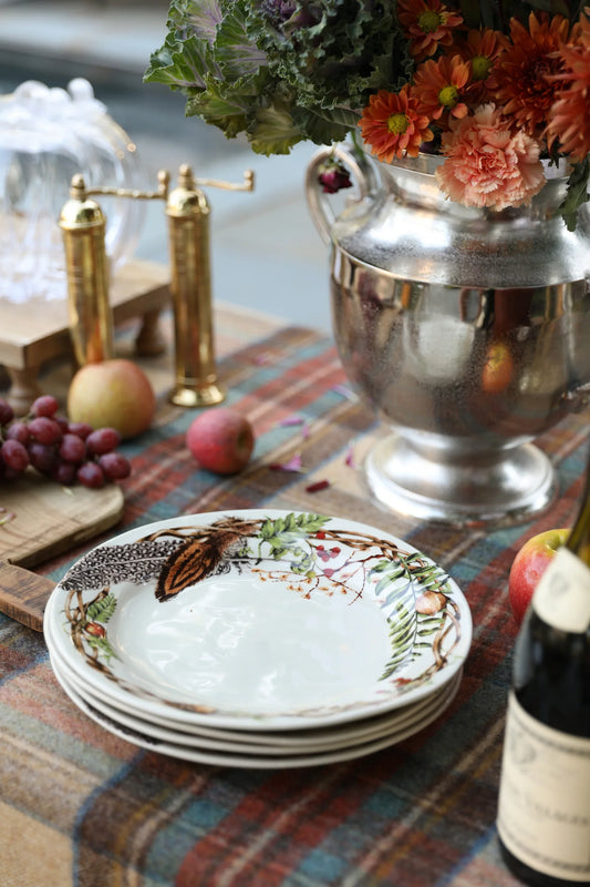Stack of decorative woodland-themed plates on a plaid tablecloth with brass pepper mills, apples, grapes, and a silver vase with autumn flowers